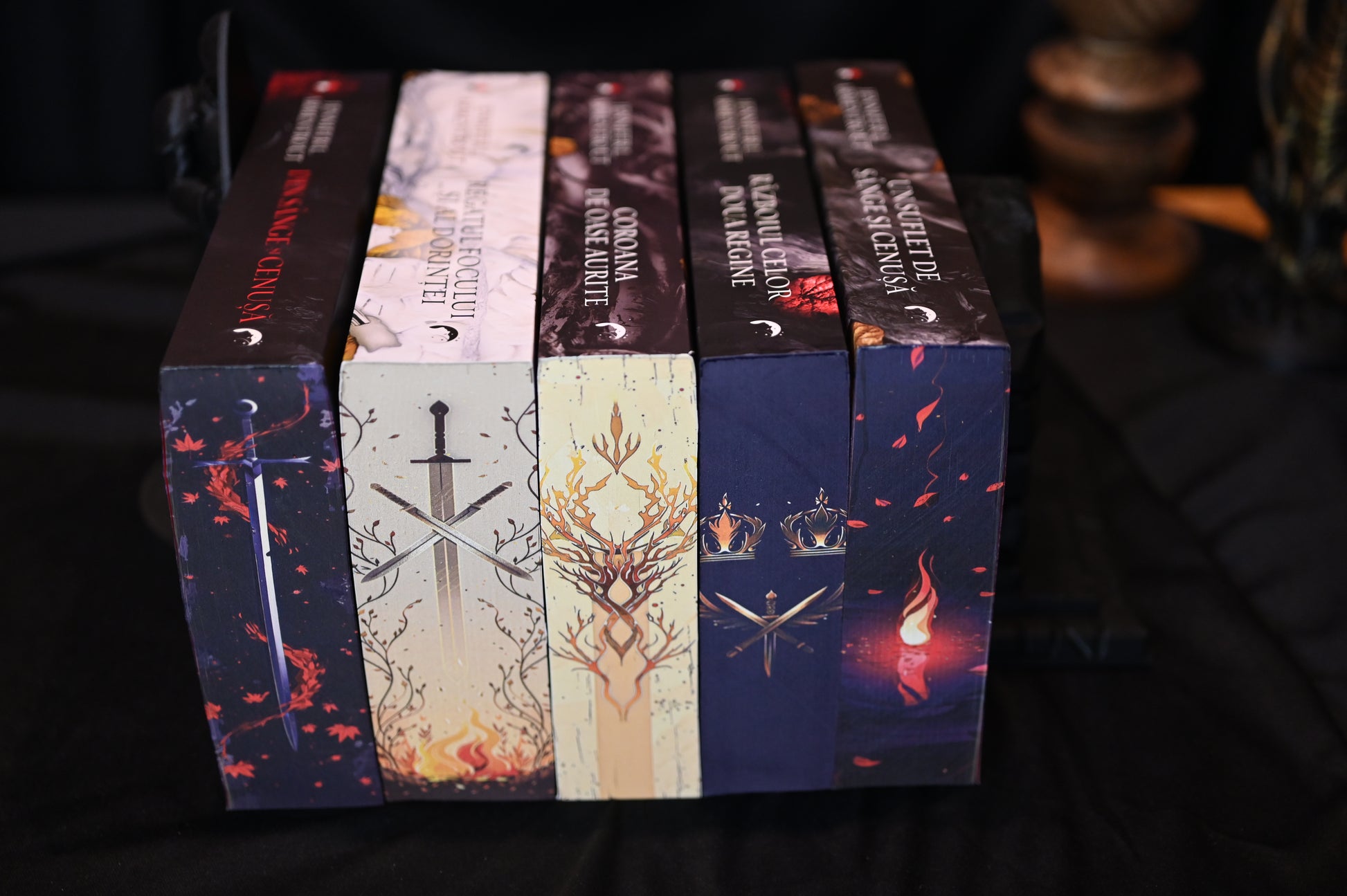 Set de cărți Din sânge și cenușă cu printed edges stilizate, prezentând designuri cu săbii încrucișate, coroane aurii, flăcări și ramuri decorative, în culori închise și tonuri calde.