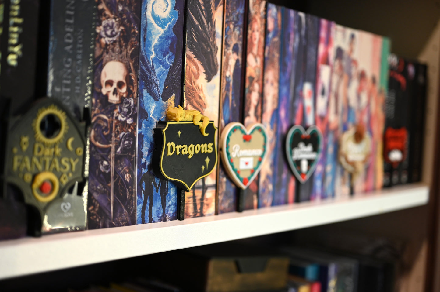 Raft de cărți decorate cu cotoare ilustrate în stil fantasy, fiecare secțiune fiind marcată cu etichete 3D tematice pentru genuri precum Dark Fantasy, Dragons, Romance, Dark Romance, Cowboy Romance și Vampires.