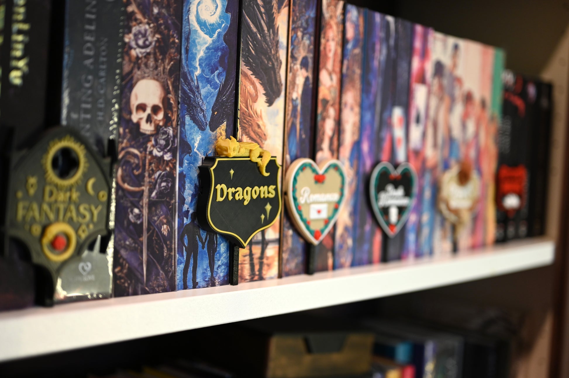Raft de cărți decorate cu cotoare ilustrate în stil fantasy, fiecare secțiune fiind marcată cu etichete 3D tematice pentru genuri precum Dark Fantasy, Dragons, Romance, Dark Romance, Cowboy Romance și Vampires.