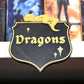 Etichetă 3D pentru secțiunea Dragons, în stil heraldic negru cu bordură aurie, decorată cu un mic dragon galben care stă întins deasupra.