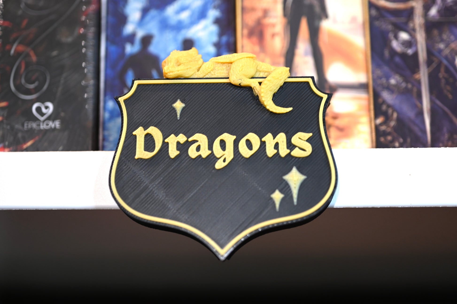 Etichetă 3D pentru secțiunea Dragons, în stil heraldic negru cu bordură aurie, decorată cu un mic dragon galben care stă întins deasupra.