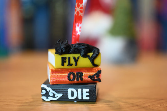 Ornament de brad inspirat din A Patra Aripă, cu un mini-dragon negru așezat pe o stivă de cărți colorate inscripționate „Fly • Or • Die”, fotografiat pe o masă din lemn cu fundal decorativ de Crăciun.