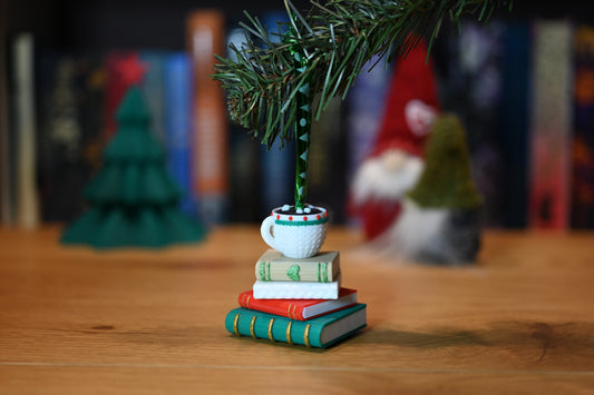 Ornament de brad cu o stivă de cărți în culori de iarnă și o cană albă cu ciocolată caldă deasupra, fotografiat pe o masă din lemn cu decor de Crăciun în fundal.