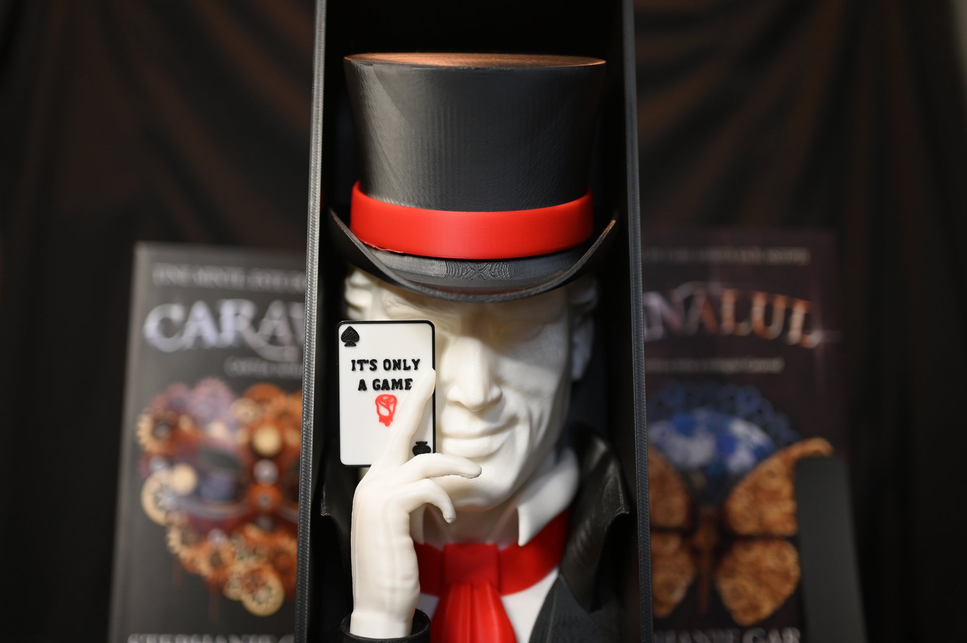 Booknook Legend - Inspirație Caraval Book nook inspirat din Legend din Caraval, încadrat între cărți cu coperți ilustrate fantasy, cu joben negru și cartelă ‘It’s Only a Game’ în mână.