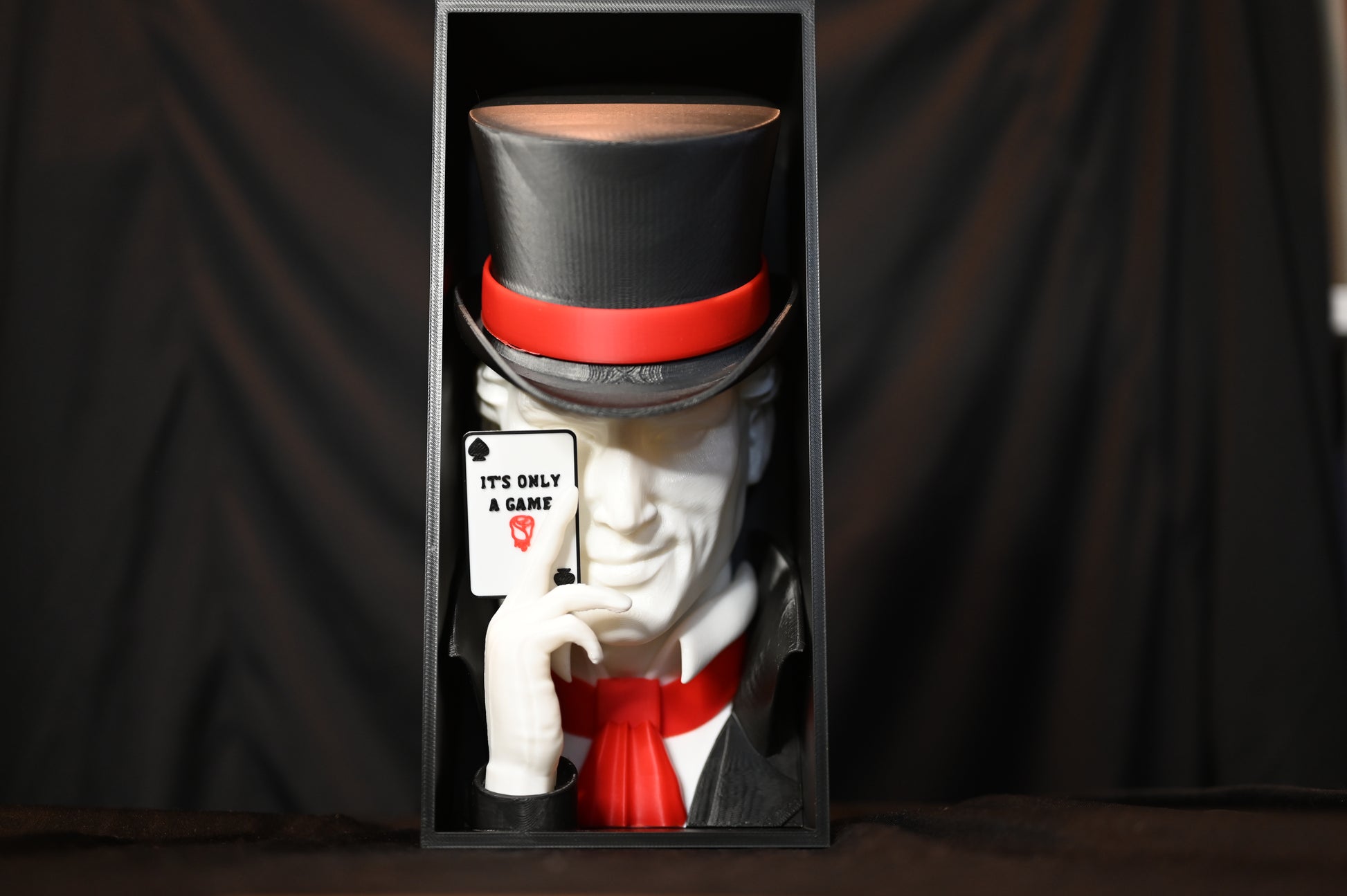 Booknook Legend - Inspirație Caraval Book nook cu figura misterioasă inspirată din Legend din Caraval, purtând joben negru cu bandă roșie și ținând o carte de joc ‘It’s Only a Game’, fotografiat pe fundal negru.