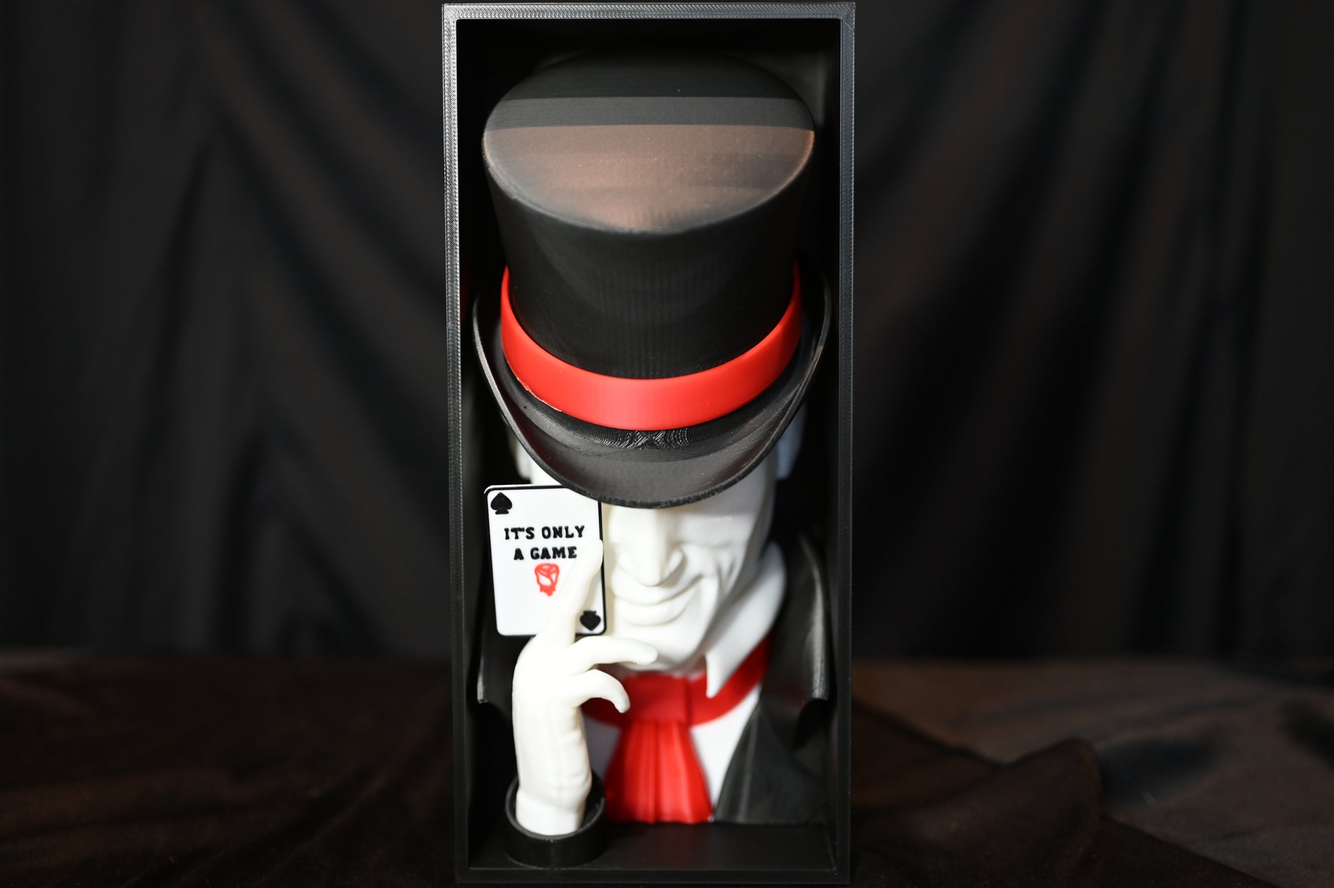 Booknook Legend - Inspirație Caraval Book nook cu figura misterioasă inspirată din Legend din Caraval, purtând joben negru cu bandă roșie și ținând o carte de joc ‘It’s Only a Game’, fotografiat pe fundal negru.