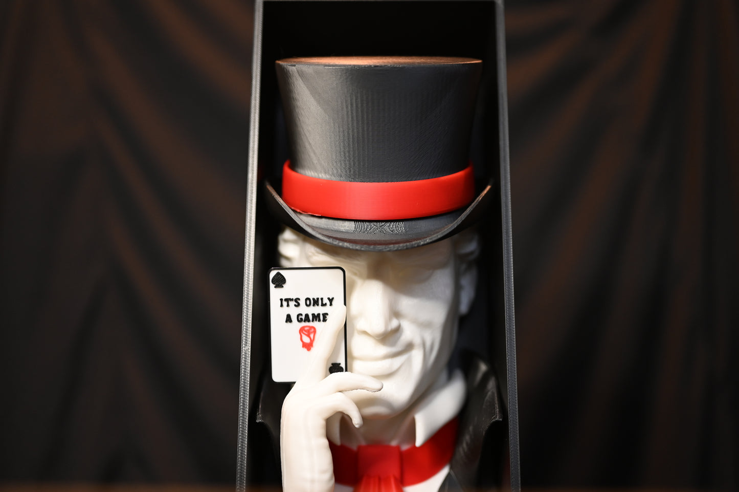 Booknook Legend - Inspirație Caraval Book nook cu figura misterioasă inspirată din Legend din Caraval, purtând joben negru cu bandă roșie și ținând o carte de joc ‘It’s Only a Game’, fotografiat pe fundal negru.