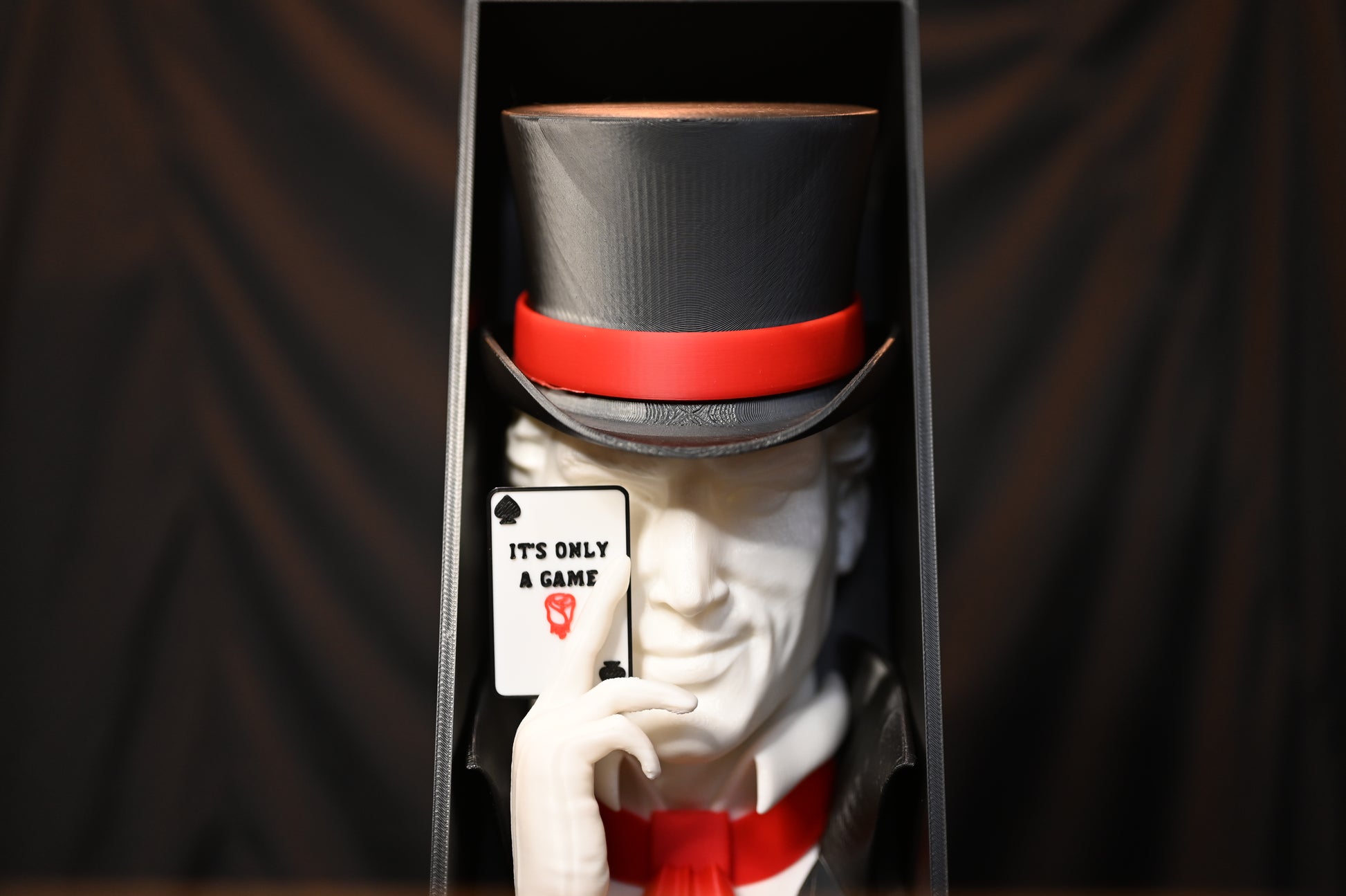 Booknook Legend - Inspirație Caraval Book nook cu figura misterioasă inspirată din Legend din Caraval, purtând joben negru cu bandă roșie și ținând o carte de joc ‘It’s Only a Game’, fotografiat pe fundal negru.