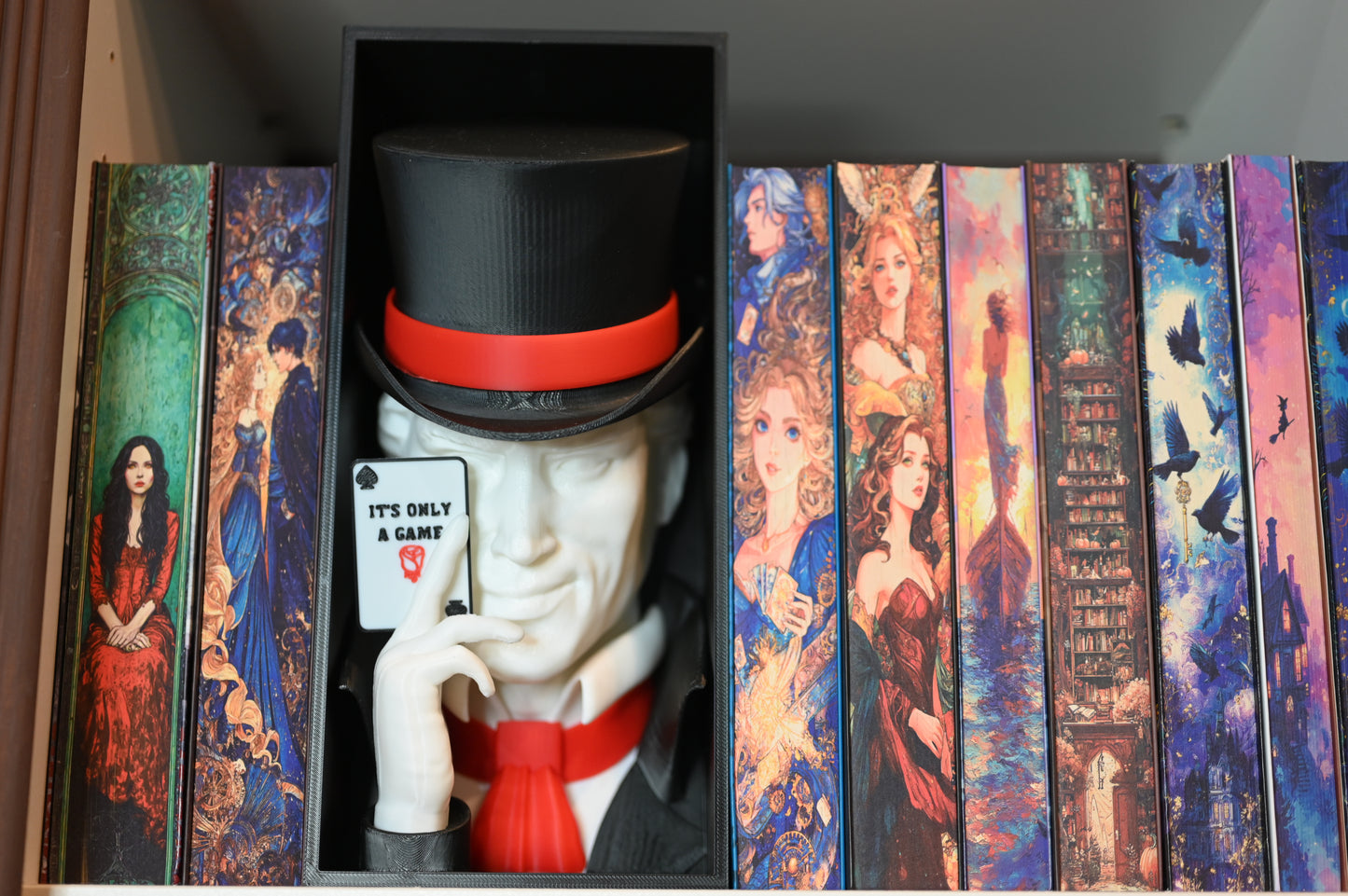 Booknook Legend - Inspirație Caraval Book nook inspirat din Legend din Caraval, încadrat între cărți cu coperți ilustrate fantasy, cu joben negru și cartelă ‘It’s Only a Game’ în mână.