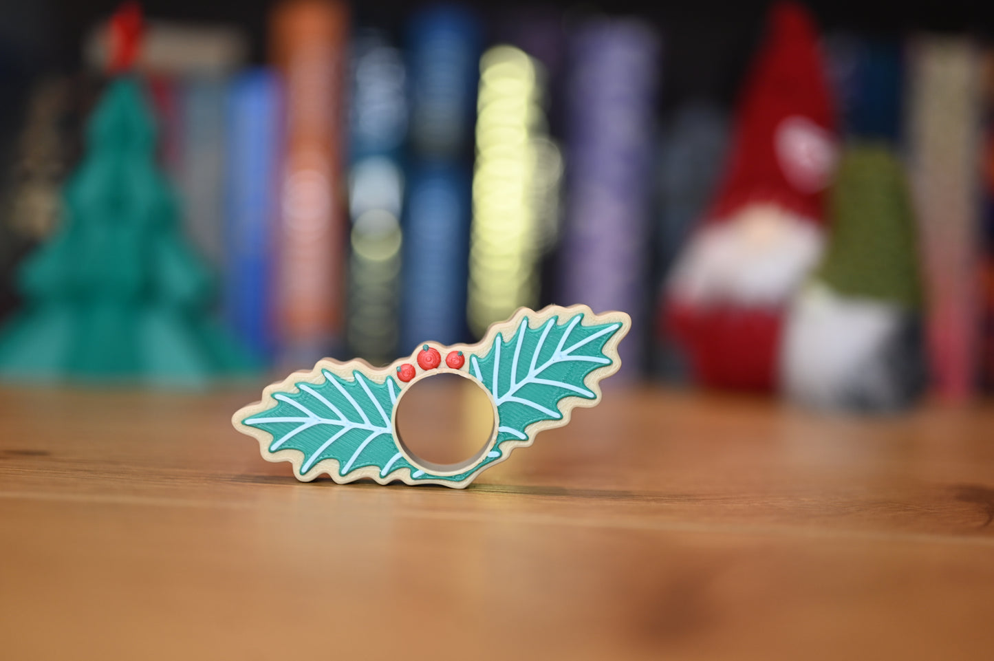 page holder decorat cu două frunze verzi de ilex și trei bobițe roșii, așezat pe o masă din lemn, cu decoruri de Crăciun neclare în fundal.