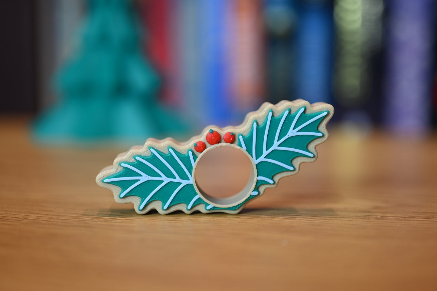 page holder decorat cu două frunze verzi de ilex și trei bobițe roșii, așezat pe o masă din lemn, cu decoruri de Crăciun neclare în fundal.