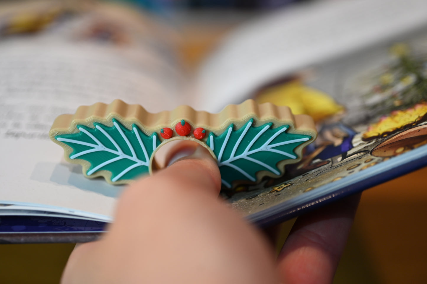 page holder decorat cu două frunze verzi de ilex și trei bobițe roșii, așezat pe o masă din lemn, cu decoruri de Crăciun neclare în fundal.