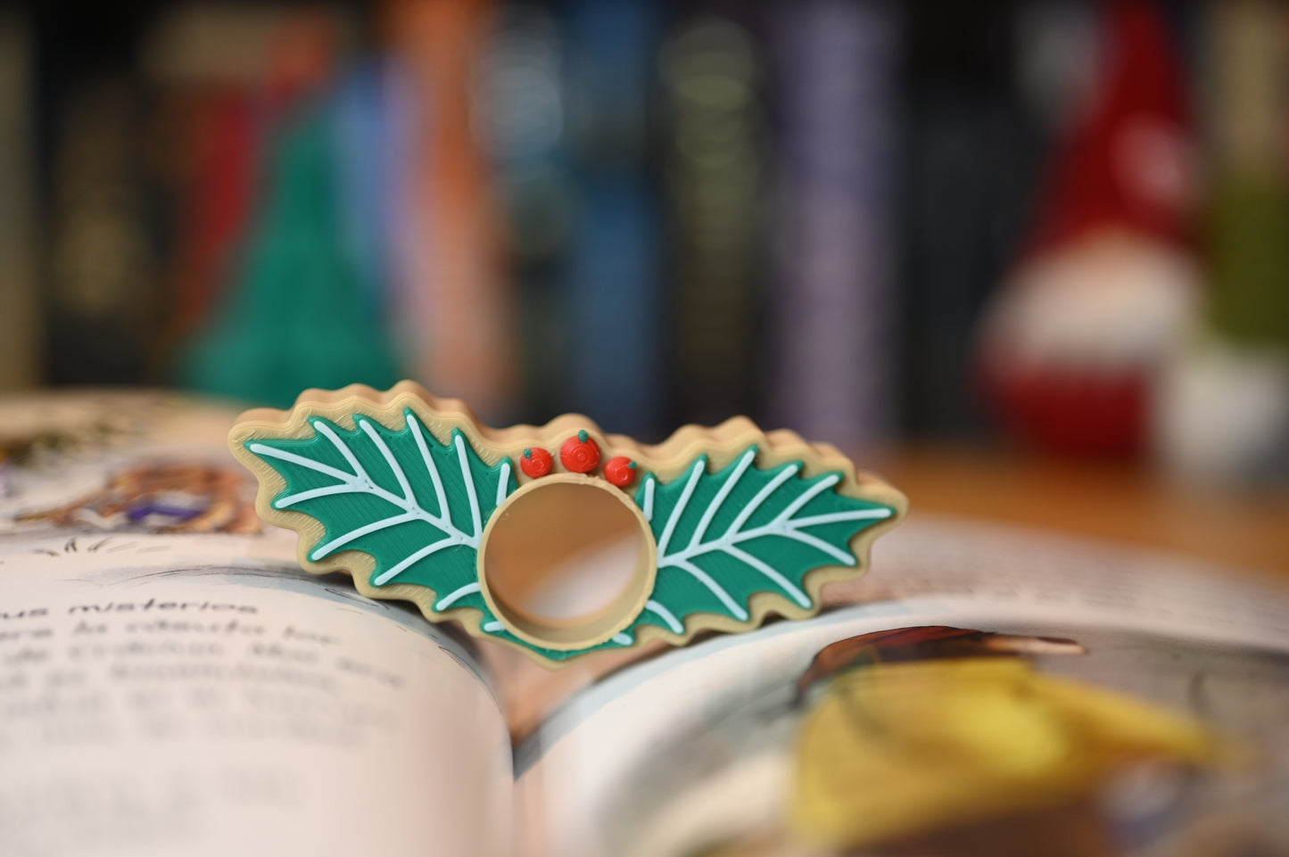 page holder decorat cu două frunze verzi de ilex și trei bobițe roșii, așezat pe o masă din lemn, cu decoruri de Crăciun neclare în fundal.