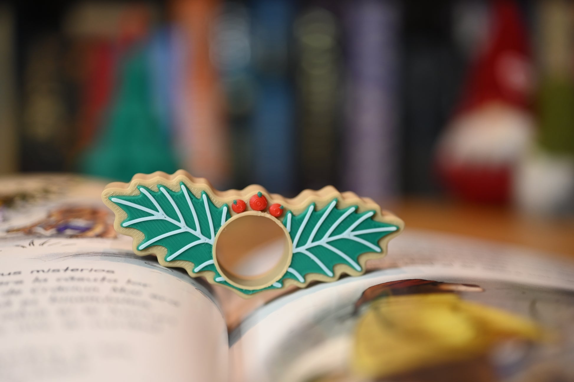 page holder decorat cu două frunze verzi de ilex și trei bobițe roșii, așezat pe o masă din lemn, cu decoruri de Crăciun neclare în fundal.