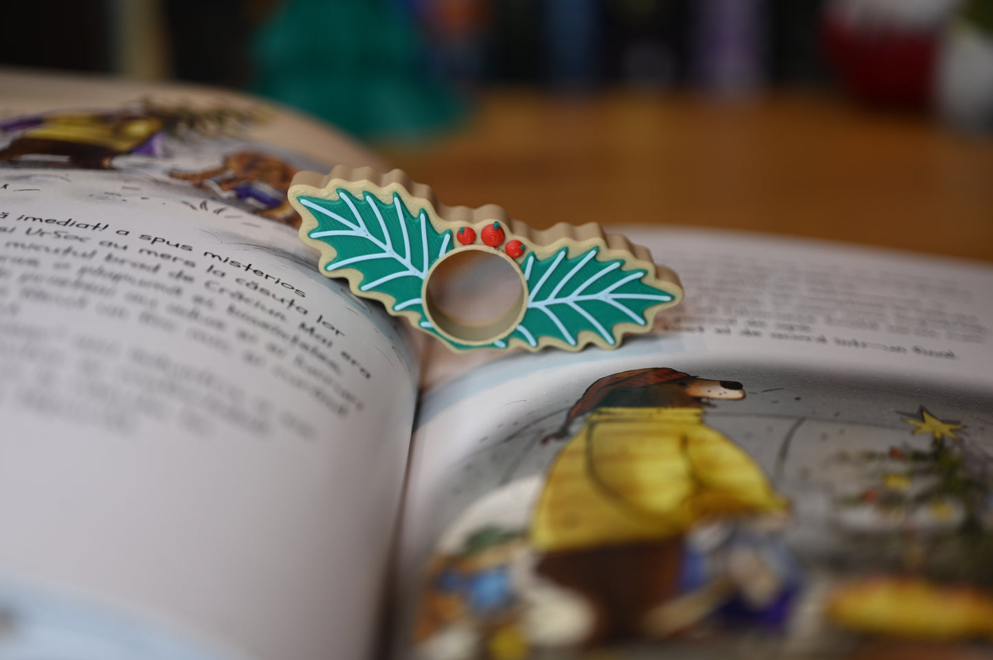 page holder decorat cu două frunze verzi de ilex și trei bobițe roșii, așezat pe o masă din lemn, cu decoruri de Crăciun neclare în fundal.