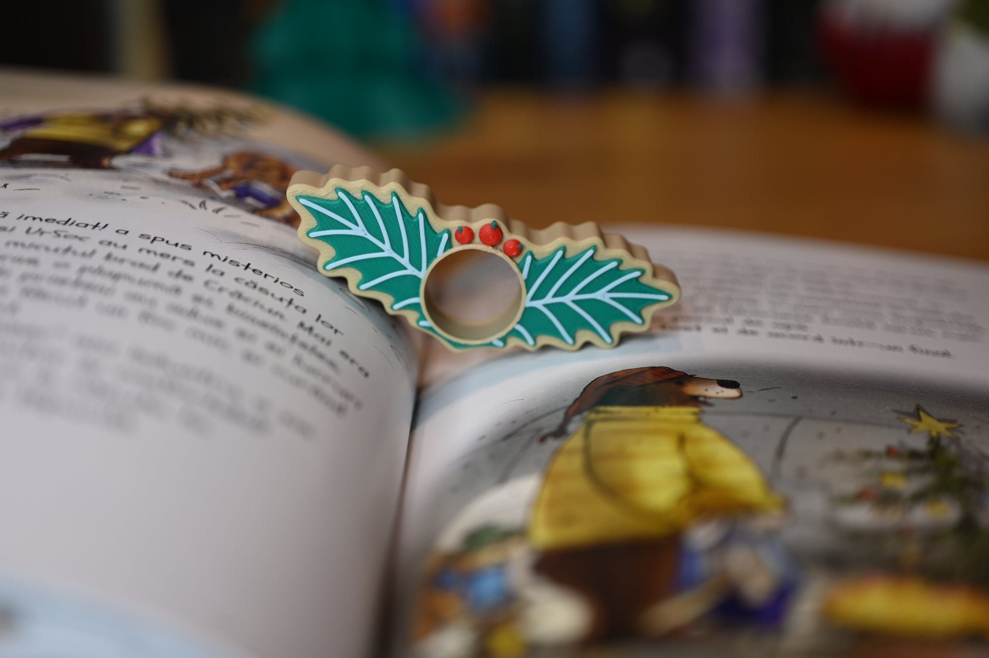 page holder decorat cu două frunze verzi de ilex și trei bobițe roșii, așezat pe o masă din lemn, cu decoruri de Crăciun neclare în fundal.