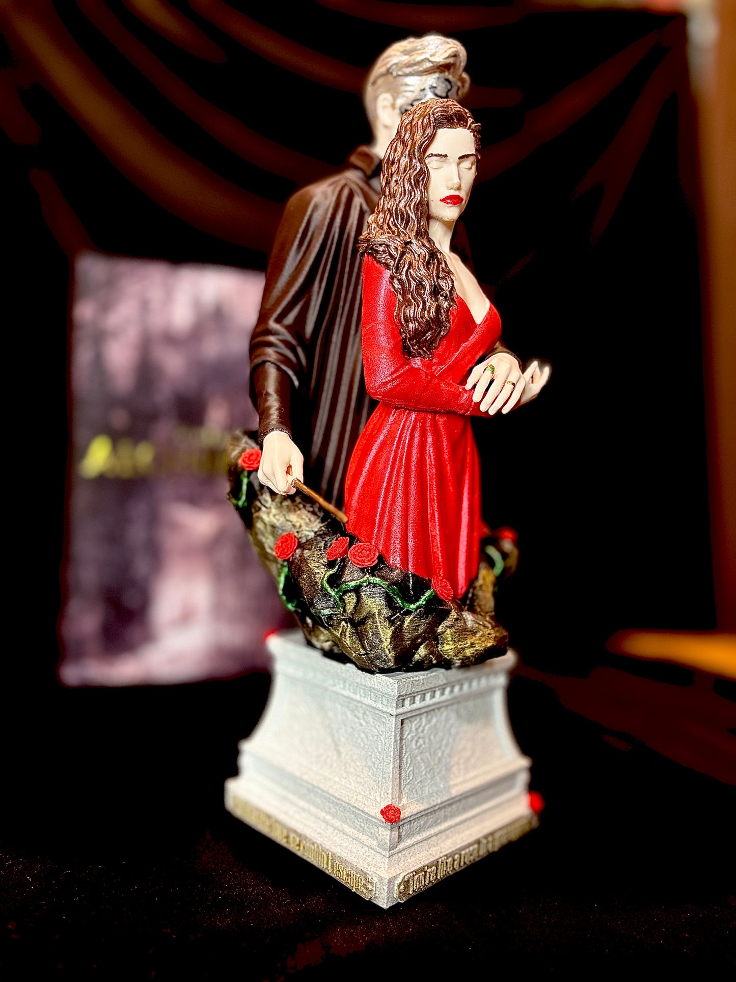 Statuetă 3D inspirată din Alchemised  O figurină decorativă fantasy ce înfățișează un bărbat cu mască metalică, îmbrăcat în negru, stând protectiv în spatele unei femei cu rochie roșie și păr ondulat. Figurină este amplasată pe un soclu ornamentat cu trandafiri roșii. Alături, se află o carte ediție specială „Alchemised”, cu o copertă gotică ilustrând un castel misterios și personajele principale în stil artistic.