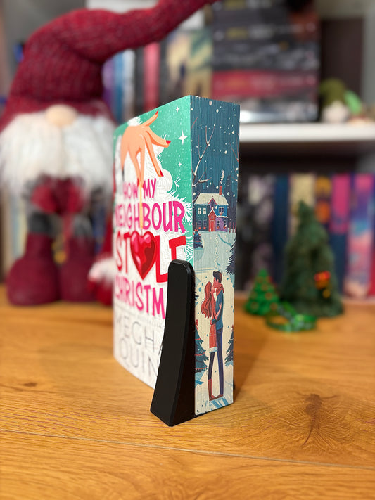 Carte intitulată ‘How My Neighbour Stole Christmas’ sprijinită într-un suport negru, cu marginile imprimate ce arată un cuplu îmbrățișat într-un peisaj de iarnă, fotografiată pe o masă de lemn cu decoruri festive în fundal.