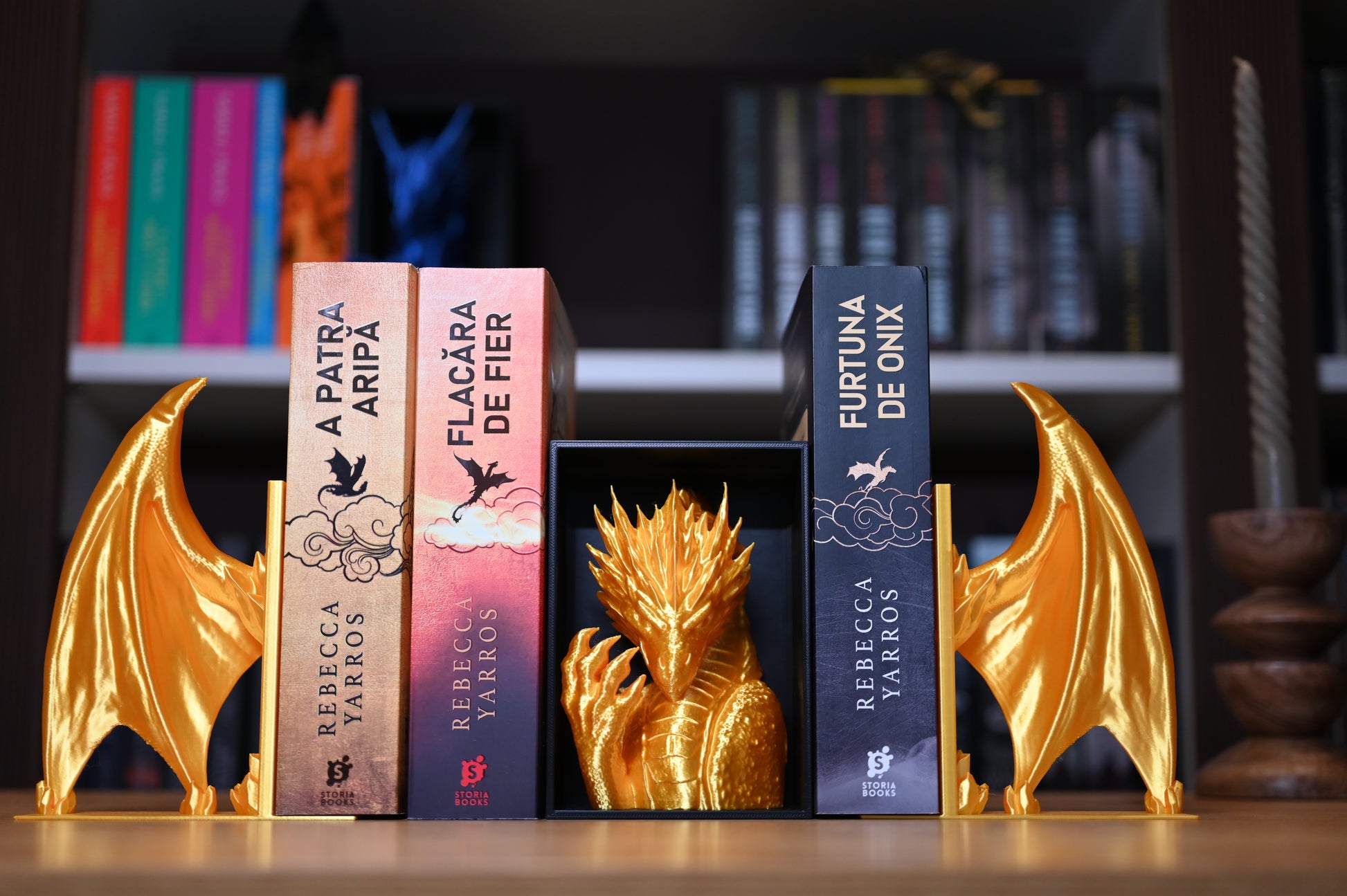 Set dioramă și suport de carte  în formă de dragon auriu, plasat între volume fantasy de Rebecca Yarros pe un raft de bibliotecă.