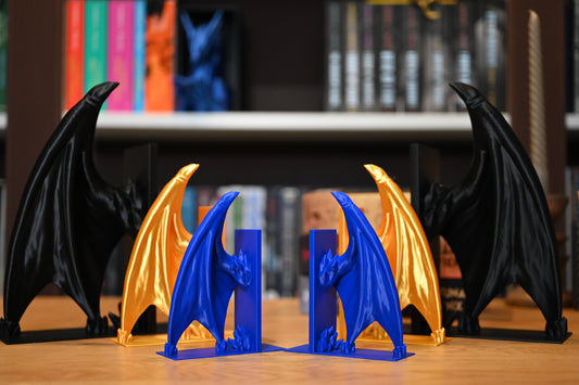 Suporturi de carte fantasy bookend in forma de aripi de dragoni, cu mai multe culori: negru, albastru, auriu, iar pe fundal sunt carti blurate