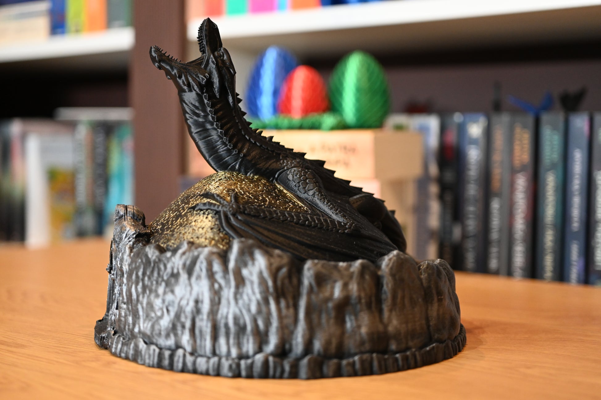 Figurină decorativă cu dragon negru pe un munte auriu, lângă ouă de dragon colorate, pe fundal cu rafturi pline de cărți fantasy.