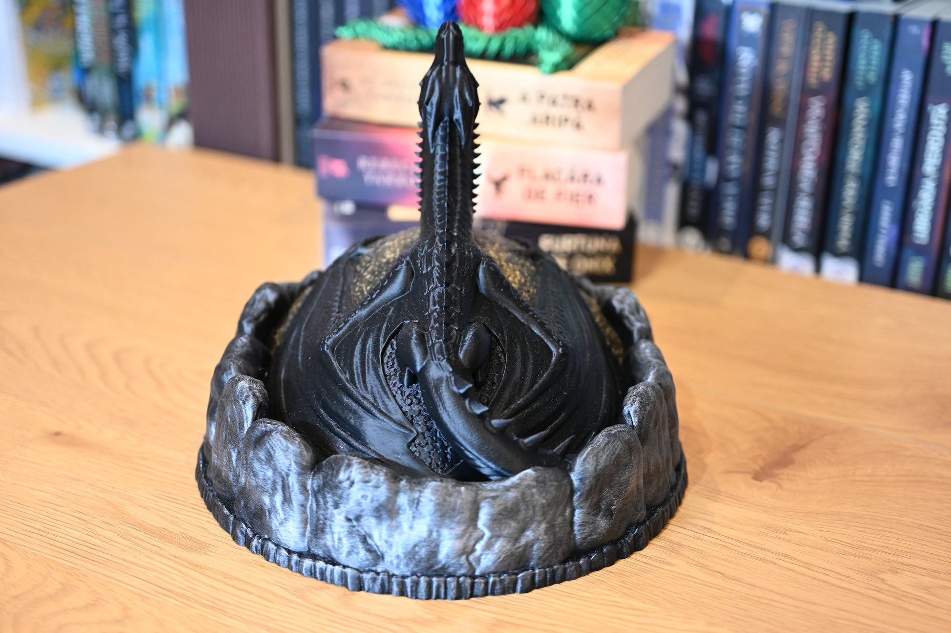 Cutia de Comori a Dragonului vedere din spate a unei figurine decorative cu dragon negru pe un munte auriu, lângă ouă de dragon colorate, pe fundal cu rafturi pline de cărți scrise de Rebecca Yarros