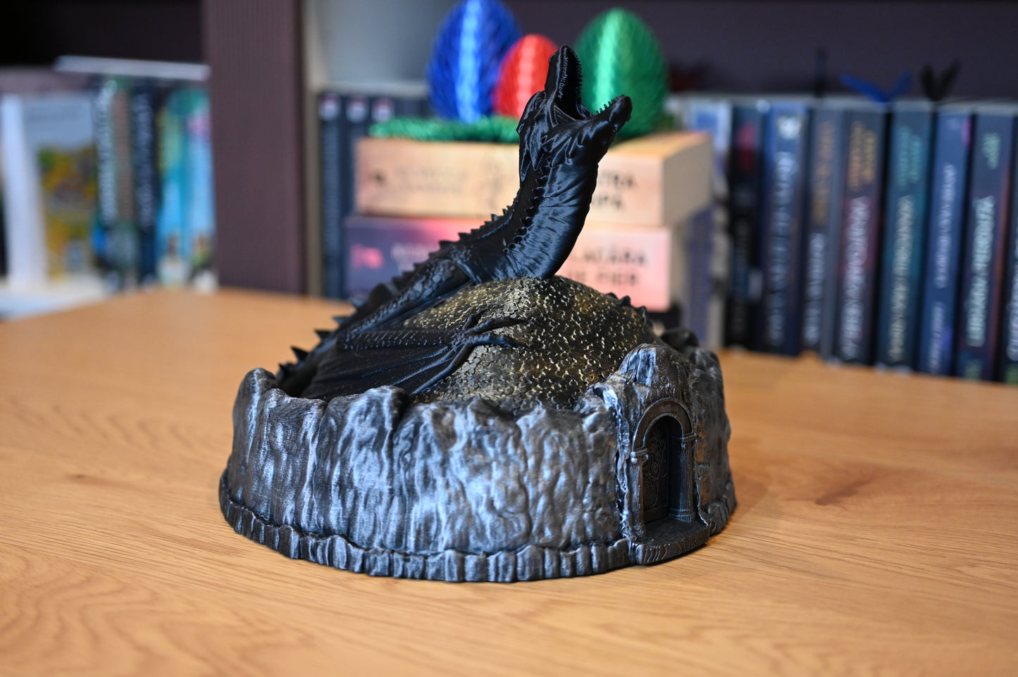 Figurină decorativă cu dragon negru pe un munte auriu, lângă ouă de dragon colorate, pe fundal cu carti din seria Empyrean