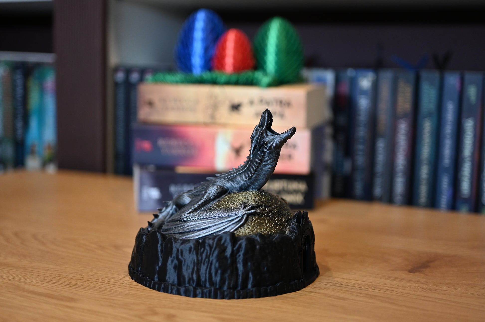 Figurină decorativă cu dragon argintiu pe un munte auriu, lângă ouă de dragon colorate, pe fundal sunt cartile seriei A patra aripa