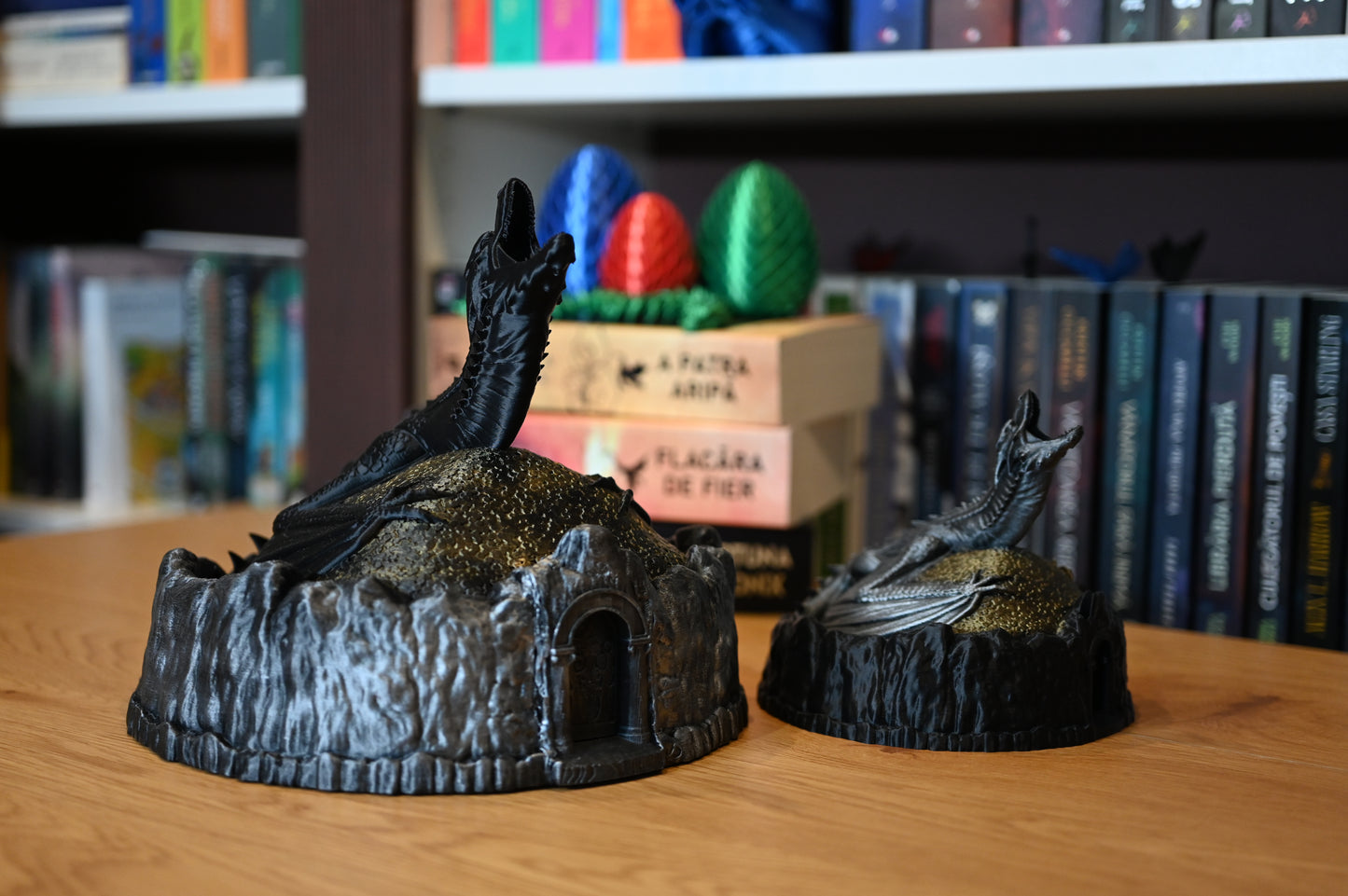doua Figurine decorative cu dragoni, unul negru, celalalt argintiu, pe un munte auriu, lângă ouă de dragon colorate, pe fundal cu rafturi pline de cărți fantasy.