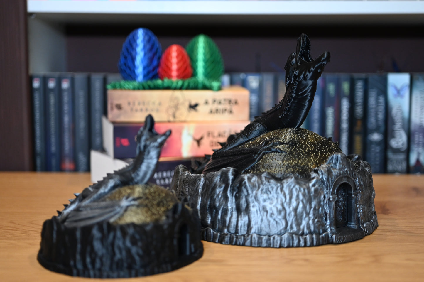 doua Figurine decorative cu dragoni, unul negru, celalalt argintiu, pe un munte auriu, lângă ouă de dragon colorate, pe fundal cu rafturi pline de cărți fantasy