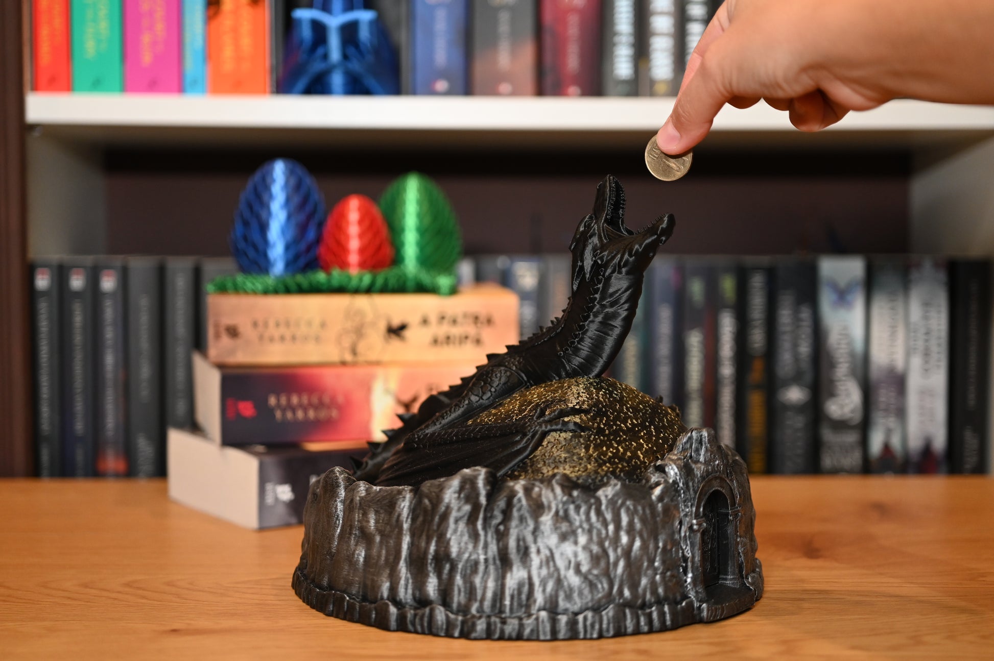 Figurina decorativa cu dragon negru,  in gura caruia poti introduce monede, pe un munte auriu, lângă ouă de dragon colorate, pe fundal cu rafturi pline de cărți fantasy.