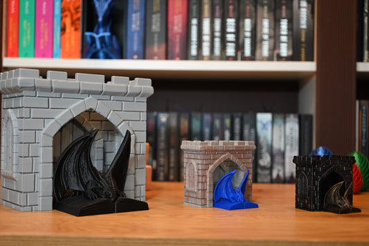 Trei figurine fantasy de dragoni în miniaturi diferite — unul negru, unul albastru și unul auriu — fiecare așezat pe o carte deschisă și încastrat într-o poartă medievală de castel, expuse pe un raft cu cărți fantasy scrise de Rebecca Yarros, Jennifer Armentrout sau Sarah  J Maas