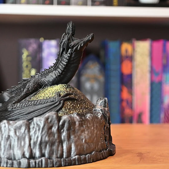 Figurină decorativă cu dragon negru pe un munte auriu, in care se introduce o moneda, pe fundal cu carti fantasy