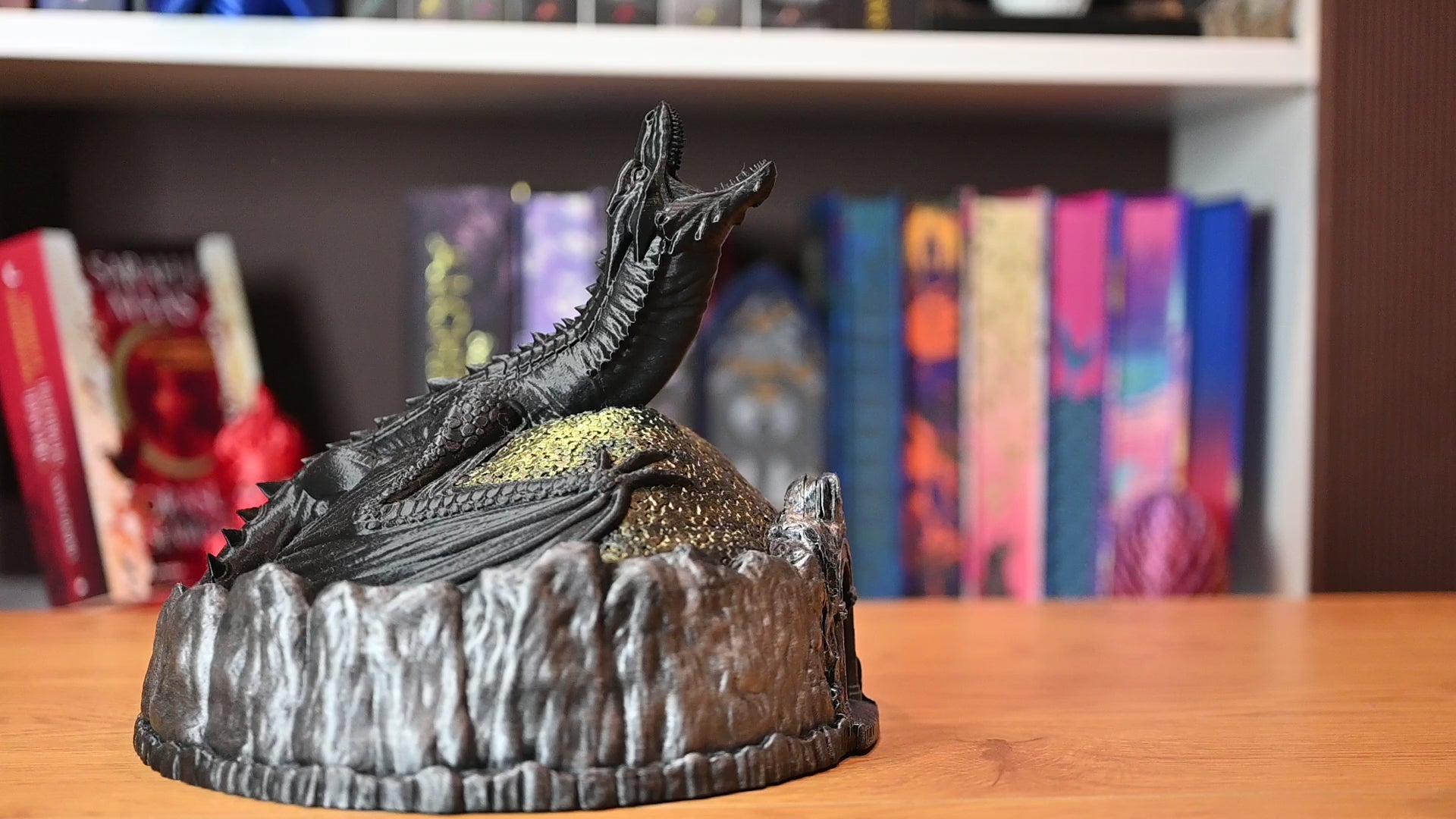 Figurină decorativă cu dragon negru pe un munte auriu, in care se introduce o moneda, pe fundal cu carti fantasy