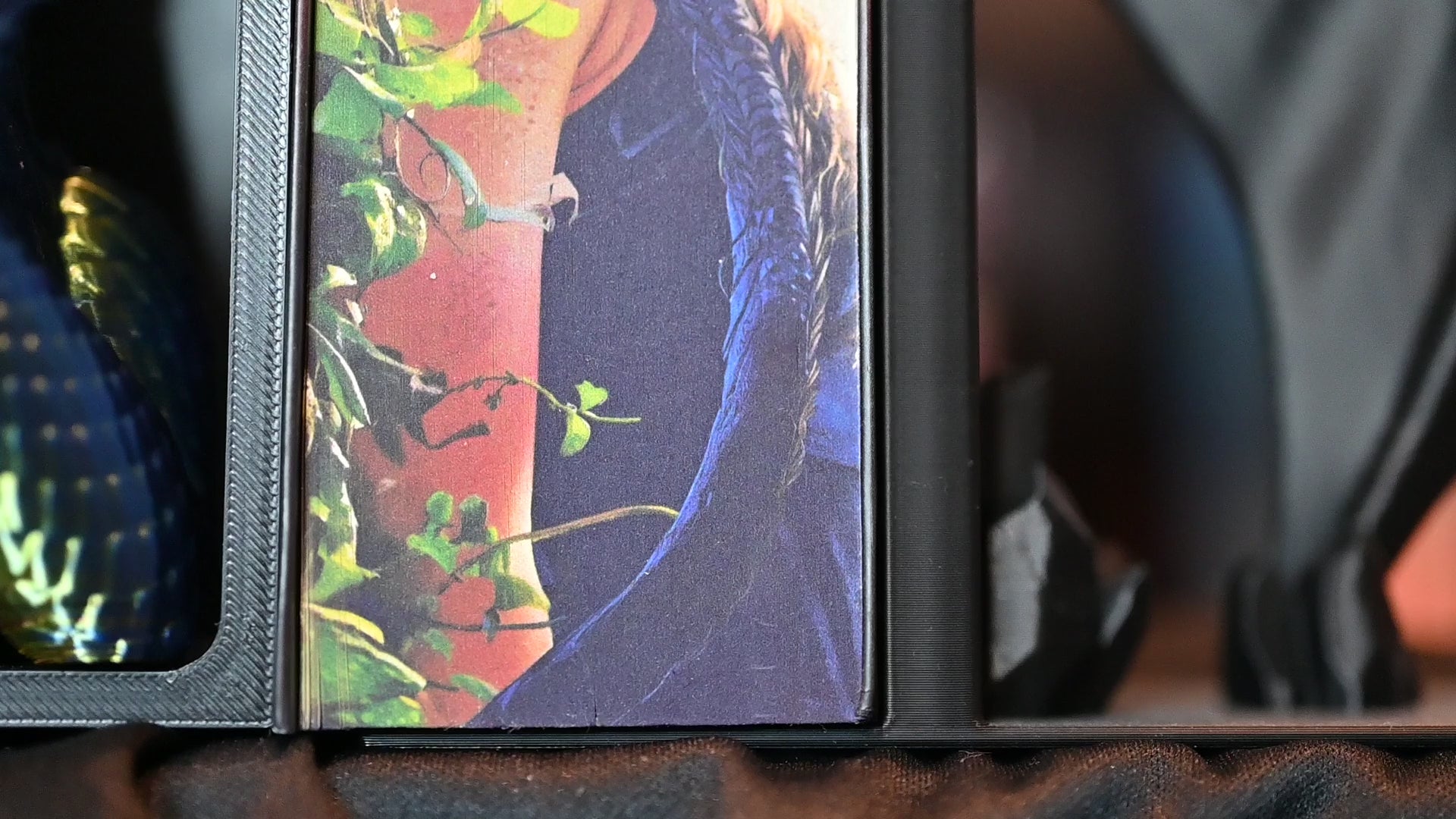 Cartea „Flacăra de fier” de Rebecca Yarros, ediție specială realizată de Printedges, prezentată între suporturi negre în formă de dragoni. Marginea imprimată (printed edge) arată portretul unei tinere înarmate și capul unui dragon negru cu ochi galbeni, ilustrând perfect universul fantasy al seriei Empyrean.